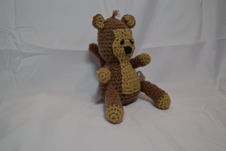 May include: A brown and tan crocheted monkey toy with black eyes. The monkey is sitting on a white surface.