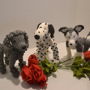 May include: Three crocheted stuffed dogs, a black and white dalmatian, a gray poodle, and a gray and white husky, are arranged on a white surface with a bouquet of red roses.