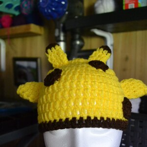 Puede incluir: Un sombrero amarillo tejido a mano con detalles marrones, dise&ntilde;ado para parecerse a una jirafa. El sombrero presenta dos peque&ntilde;as orejas y dos manchas marrones en la parte superior, con un borde marr&oacute;n en la base. Este accesorio es perfecto para ni&ntilde;os.