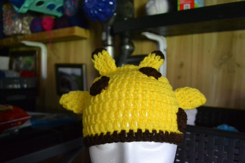 Puede incluir: Un sombrero amarillo tejido a mano con detalles marrones, dise&ntilde;ado para parecerse a una jirafa. El sombrero presenta dos peque&ntilde;as orejas y dos manchas marrones en la parte superior, con un borde marr&oacute;n en la base. Este accesorio es perfecto para ni&ntilde;os.