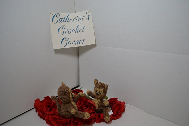 May include: Two brown crocheted monkey toys sitting on a bed of red fabric roses. The sign above reads "Catherine's Crochet Corner."