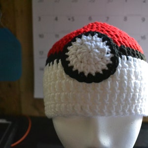 Puede incluir: Un gorro de ganchillo hecho a mano al estilo de una Pok&eacute; Ball. El gorro presenta una parte superior roja, un centro blanco y una banda negra. Un dise&ntilde;o circular blanco y negro est&aacute; centrado en la parte blanca del gorro. El gorro se exhibe en una cabeza de maniqu&iacute; blanca.