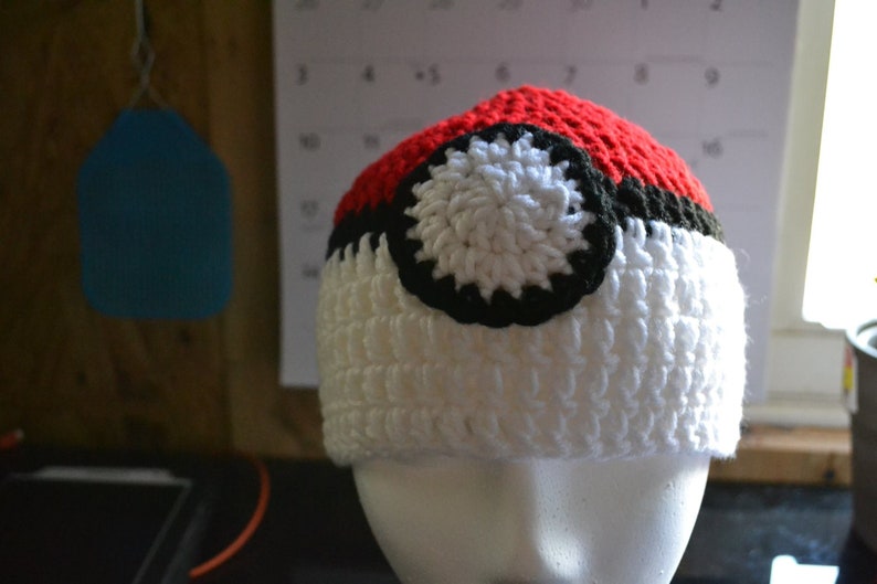 Puede incluir: Un gorro de ganchillo hecho a mano al estilo de una Pok&eacute; Ball. El gorro presenta una parte superior roja, un centro blanco y una banda negra. Un dise&ntilde;o circular blanco y negro est&aacute; centrado en la parte blanca del gorro. El gorro se exhibe en una cabeza de maniqu&iacute; blanca.