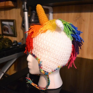 Puede incluir: Un gorro de unicornio blanco tejido a ganchillo con un cuerno amarillo, orejas blancas y una melena arco&iacute;ris. El gorro est&aacute; dise&ntilde;ado para la cabeza de un ni&ntilde;o y tiene una cinta trenzada. El gorro se muestra en una cabeza de maniqu&iacute; blanca.