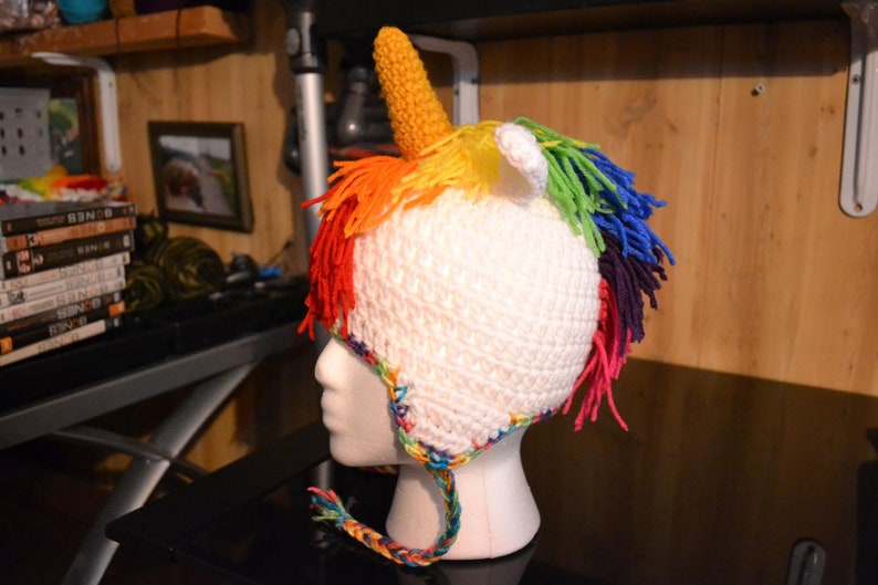 Puede incluir: Un gorro de unicornio blanco tejido a ganchillo con un cuerno amarillo, orejas blancas y una melena arco&iacute;ris. El gorro est&aacute; dise&ntilde;ado para la cabeza de un ni&ntilde;o y tiene una cinta trenzada. El gorro se muestra en una cabeza de maniqu&iacute; blanca.