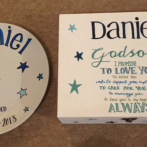 Puede incluir: Un plato de cerámica blanco con el nombre "Daniel" escrito en letras cursivas azules y estrellas azules. El plato también dice "Bautizado el 25 de noviembre de 2018". Una caja de madera blanca con un diseño de estrellas azules y el texto "Daniel Ahijado Prometo amarte guiarte apoyarte cuidarte animarte tenerte en mi corazón SIEMPRE".