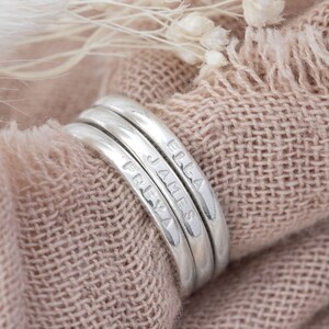 May include: Three silver rings, each inscribed with a name: FREYA, JAMES, and ELLA. The rings are stacked and presented on a peach-coloured fabric, with dried flowers in the background. The rings have a simple, elegant design.