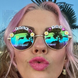 May include: Round sunglasses with gold frames and mirrored lenses reflecting a tropical scene. The top of the frames feature the words "FUCK YOU" spelled out with letter beads, and are decorated with small white flowers.