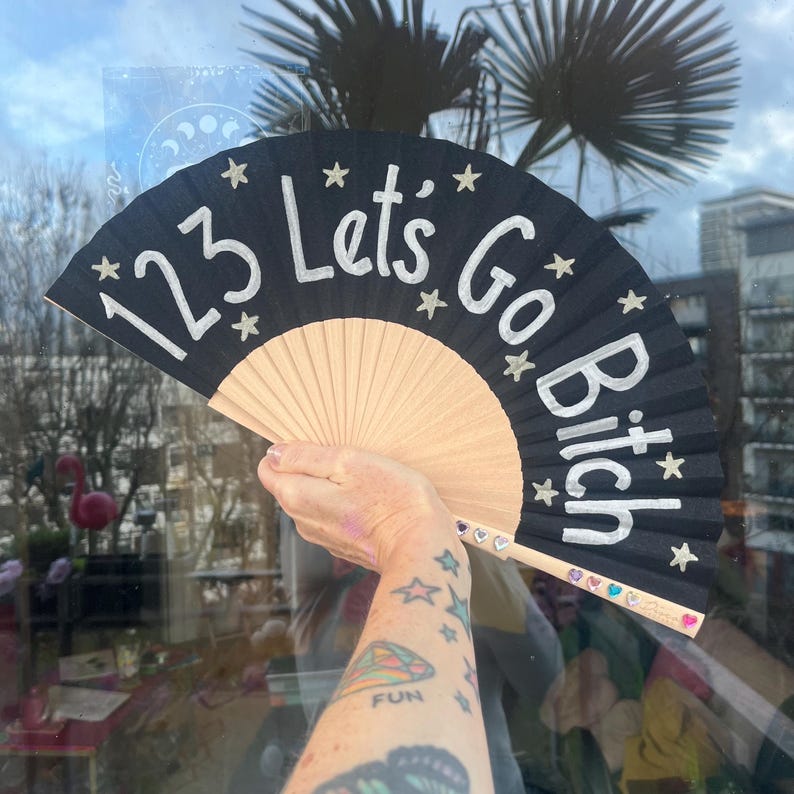 May include: Black hand fan with the text "123 Lets Go Bitch" in white, accented with stars. The fan has a natural wood-colored base and is held up by a person.