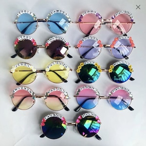 May include: A set of 12 pairs of round sunglasses with different coloured lenses and decorative text on the frames. The text includes words like "Veggie", "Samosa", "Jerry", "Palozza", "Burger", "Beer", "Beef", "Wellie", "Chicken", "Waffle", "Caesar", and "Salad".
