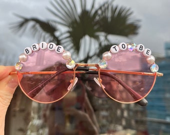 May include: Pink round sunglasses with gold frames and white beads spelling "BRIDE TO BE" on the top of the frames.