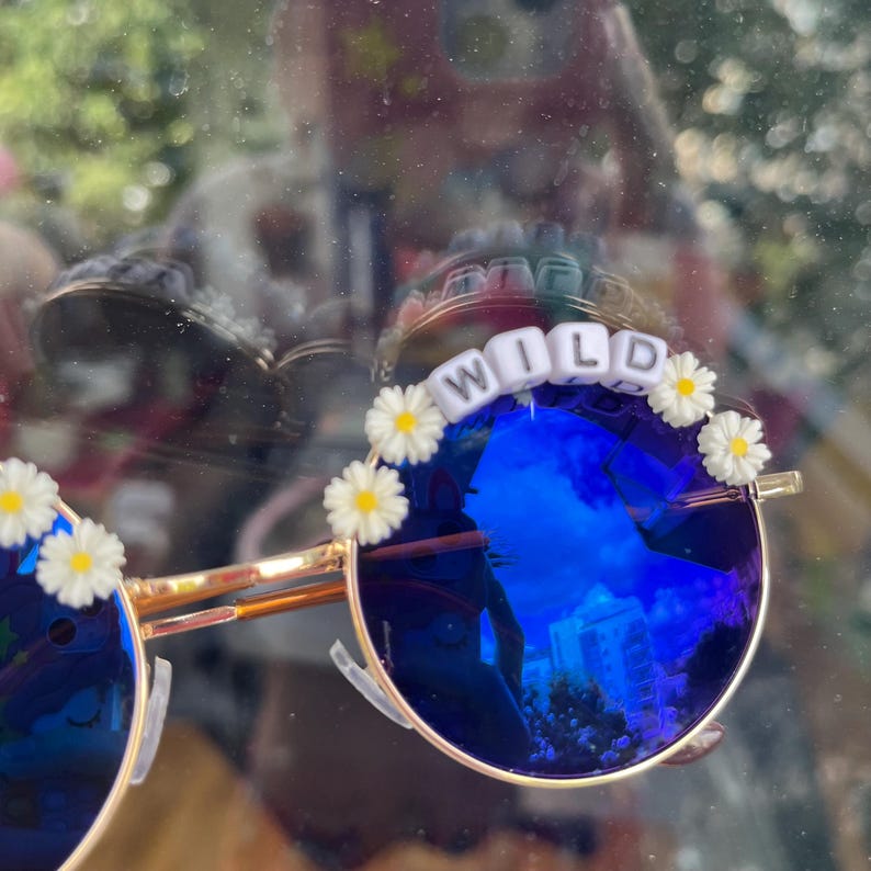 Puede incluir: Gafas de sol redondas con montura dorada y lentes azules espejadas. Las gafas tienen adornos blancos en forma de margarita y cuentas de letras blancas que deletrean "WILD" en el marco.