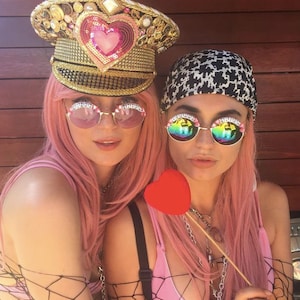 Puede incluir: Dos mujeres con pelucas rosas y gafas de sol. Una mujer lleva un sombrero adornado con lentejuelas doradas y rosas. La otra mujer lleva un pa&ntilde;uelo negro y blanco. Est&aacute;n sosteniendo un objeto rojo en forma de coraz&oacute;n.