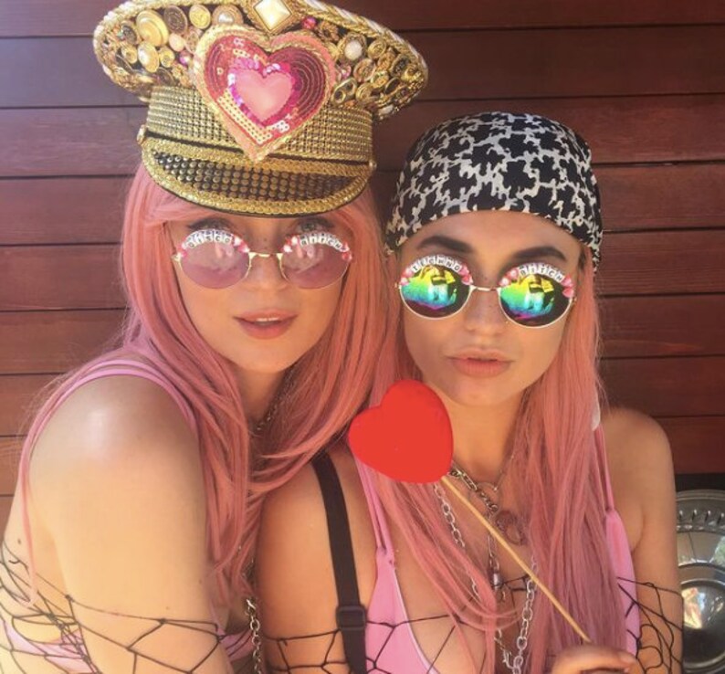 Puede incluir: Dos mujeres con pelucas rosas y gafas de sol. Una mujer lleva un sombrero adornado con lentejuelas doradas y rosas. La otra mujer lleva un pa&ntilde;uelo negro y blanco. Est&aacute;n sosteniendo un objeto rojo en forma de coraz&oacute;n.