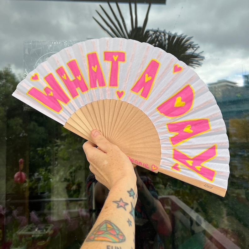 May include: A hand fan with a wooden handle and white paper. The fan has the words "WHAT A DAY" in pink letters with yellow heart accents. The fan is open, displaying its full design.