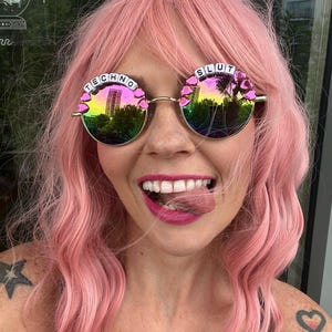 Op de afbeelding: Een vrouw met roze haar draagt ronde zonnebrillen met de woorden "TECHNO" en "SLUT" in witte letters op de monturen.