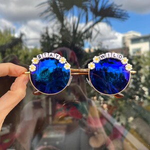 Puede incluir: Gafas de sol redondas azules con monturas doradas y detalles de margaritas blancas. Las gafas de sol tienen las palabras "STAY WILD" escritas con cuentas blancas en las monturas.