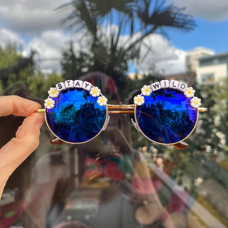 Puede incluir: Gafas de sol redondas azules con monturas doradas y detalles de margaritas blancas. Las gafas de sol tienen las palabras "STAY WILD" escritas con cuentas blancas en las monturas.