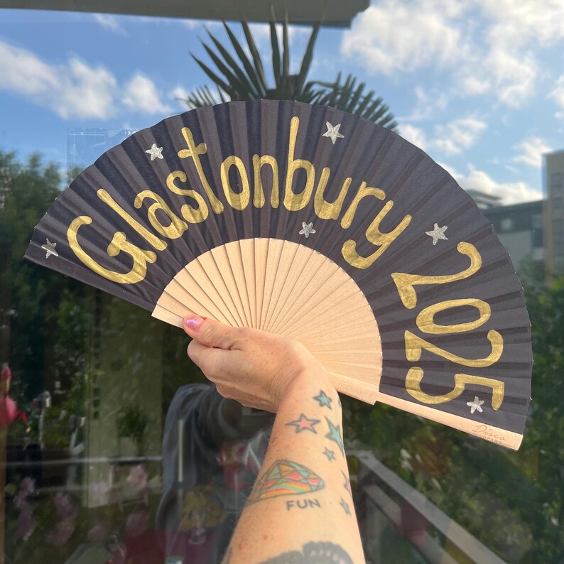 May include: A black hand fan with the gold text "Glastonbury 2025" and star accents. The fan has a natural wood base and is held up against a window with a blue sky background.