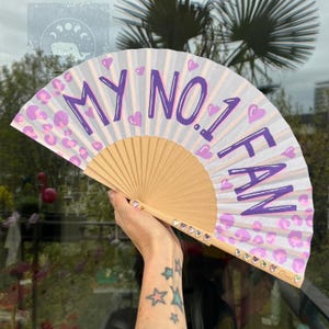 May include: A hand fan with a wooden base and white paper. The fan has the text "MY NO.1 FAN" in purple, along with purple hearts and leopard print accents. The fan is open, displaying its full design.