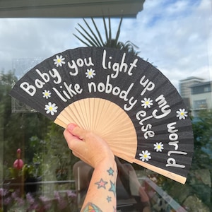 May include: A black hand fan with the text "Baby you light up my world like nobody else" in white script. The fan features daisy accents and a natural wood handle.
