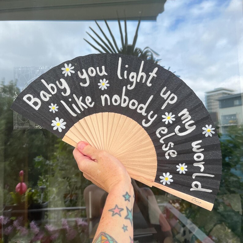 May include: A black hand fan with the text "Baby you light up my world like nobody else" in white script. The fan features daisy accents and a natural wood handle.