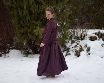 May include: A woman wearing a long, dark purple skirt and a matching long-sleeved top stands in a snowy landscape. She is barefoot and smiling.