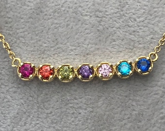 Puede incluir: Un collar de cadena de oro con una fila de piedras preciosas de colores. Las piedras preciosas son rojas, naranjas, amarillas, verdes, moradas, rosas, azules y azules.