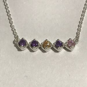 Puede incluir: Un collar de cadena de plata con cinco piedras preciosas. Las piedras preciosas son moradas, moradas, doradas, moradas y rosadas.