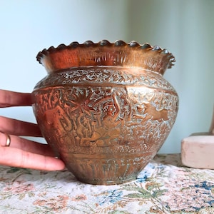 May include: A vintage copper-coloured vase with detailed etched patterns. The vase has a broad, rounded form and a frilled rim. The etched designs show figures and animals, enhancing the vessel's decorative appeal. The vase is likely for ornamental use.