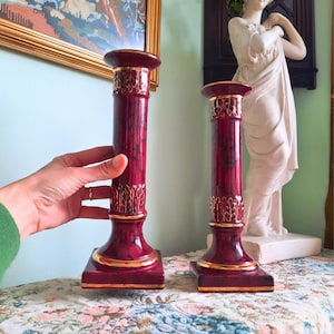 May include: A pair of burgundy candle holders with gold detailing. The candle holders have a column design with a square base and a round top. Gold accents are on the top, bottom, and middle of the column. They are displayed on a floral patterned surface.