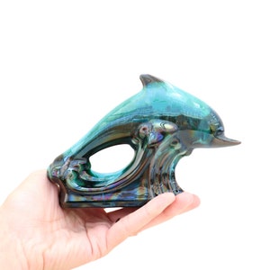 May include: A ceramic dolphin figurine with a teal and iridescent glaze. The dolphin is leaping out of the water, which is depicted as a wave.