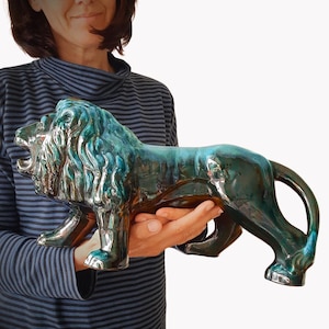 Könnte beinhalten: Eine Keramik-Löwenfigur mit einer glänzenden, mehrfarbigen Glasur in Blaugrün und Dunkelblau. Der Löwe ist in stehender Seitenansicht mit geöffnetem Maul dargestellt und zeigt detaillierte, modellierte Fellstrukturen an Mähne und Körper.