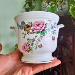 May include: White ceramic planter with pink floral and fruit basket design. The planter has two small handles and a gold trim around the base. The floral design includes pink peonies and green leaves.