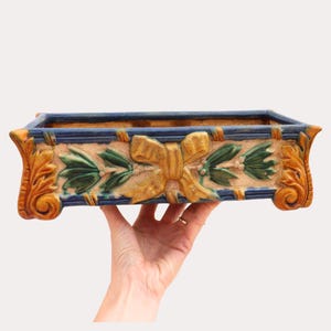 Könnte beinhalten: Rechteckiger Keramik-Pflanztopf mit blauem Rand und beigem Körper. Der Pflanztopf hat ein dekoratives Design mit einer goldenen Schleife, grünen Blättern und orangefarbenen Akzenten. Der Pflanztopf wird von einer Hand gehalten.