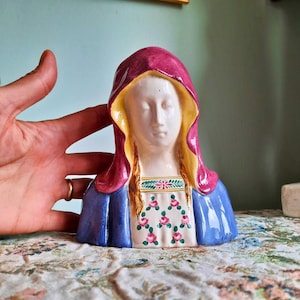 May include: A ceramic bust of a figure wearing a magenta head covering with a yellow interior lining. The figure has a white face, blonde hair, and a blue garment featuring a white chest panel decorated with a pattern of small pink roses and green leaves.