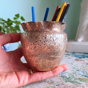 Könnte beinhalten: Ein kleiner Messingtopf im antiken Stil mit einem aufwendigen geätzten Design von Vögeln und Blättern. Der Topf hat einen gerüschten Rand und ist mit farbigen Bleistiften gefüllt. Der Topf wird in einer Hand gehalten.
