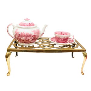 May include: A brass tea kettle stand with ornate detailing and four legs. A pink and white teapot and matching teacup and saucer are on top of the stand.