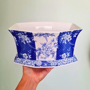 May include: A ceramic planter featuring a white base with a blue floral pattern and a checkered design. The vessel has a wide, flared rim and a tapered body with a rectangular base, showcasing intricate botanical illustrations in deep blue ink.