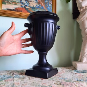 May include: A black urn with a square base, a fluted body, and two small handles. The urn is made of a matte material and sits on a floral patterned surface. The background includes a framed painting and a white statue.