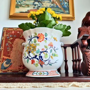 May include: A white ceramic plant pot with a floral pattern in orange, yellow, and blue. The pot contains a plant with yellow flowers and green leaves. The pot sits on a dark wooden surface.