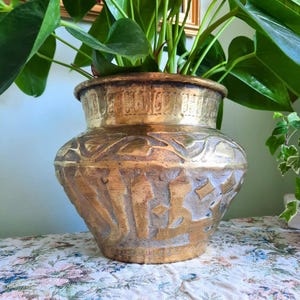 May include: A vintage brass planter with an embossed design. The planter has a wide, rounded body and a slightly flared rim. The surface is detailed with intricate patterns and textures, perfect for displaying plants.