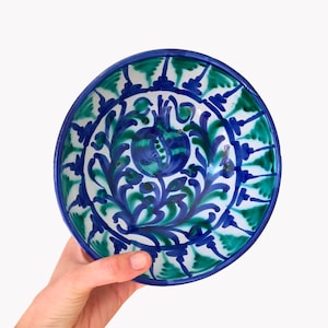 Könnte beinhalten: Handbemalte Keramikschale mit blauem Rand und einem zentralen Design mit einer blau-grünen Granatapfel. Das Innere der Schale ist mit einem sich wiederholenden Muster aus blauen und grünen Blättern und floralen Elementen verziert.
