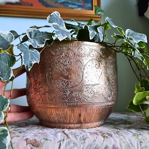 May include: A round copper planter featuring intricate embossed patterns of animals and scrolling foliage. The metallic surface displays a warm, reflective finish with a textured border at the base, containing a trailing variegated ivy plant.