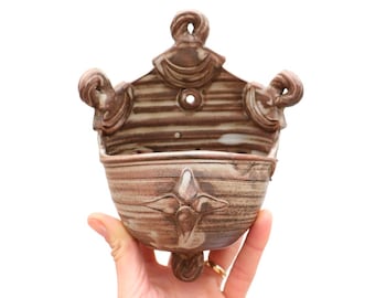 May include: A brown and white ceramic wall planter with a unique design. The planter has a rounded bowl shape with a decorative rim and a hole in the back for hanging.