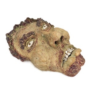 Könnte beinhalten: Eine Keramikskulptur eines Gesichts mit einem grotesken Ausdruck. Das Gesicht ist in Braun-, Grün- und Rottönen bemalt. Die Skulptur ist so gestaltet, dass sie einem Menschen mit einer großen Nase und einem breiten Mund ähnelt.