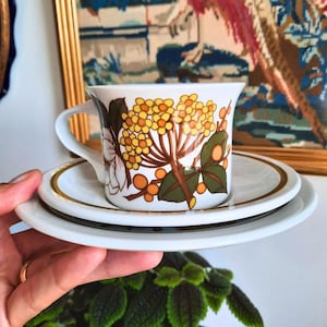 May include: A white teacup and saucer set with a floral design. The cup features a pattern of yellow and orange flowers with brown stems and green leaves. The saucer has a gold rim.