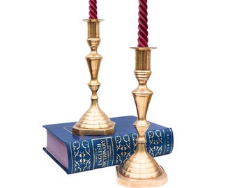 May include: Two gold-coloured brass candlesticks with burgundy spiral candles. The candlesticks are on a blue book with gold lettering. The candlesticks have a classic design, suitable for home decor, and are made of brass.
