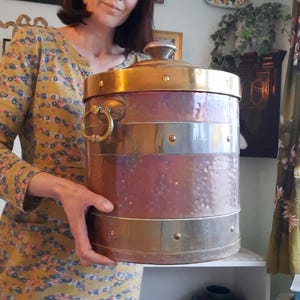 May include: A vintage copper and brass container with a lid and a handle. The container has a cylindrical shape with horizontal bands of brass and copper. The lid is brass and has a small handle on top. The container is held by a person.