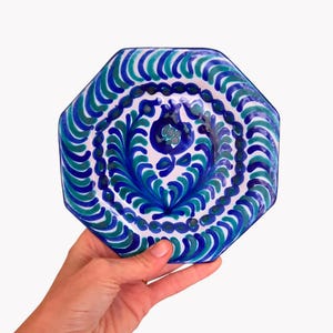 May include: An octagonal ceramic plate with a hand-painted design. The plate features a white base with a central floral motif in shades of blue and teal. The rim is decorated with a repeating pattern of blue and teal arches.